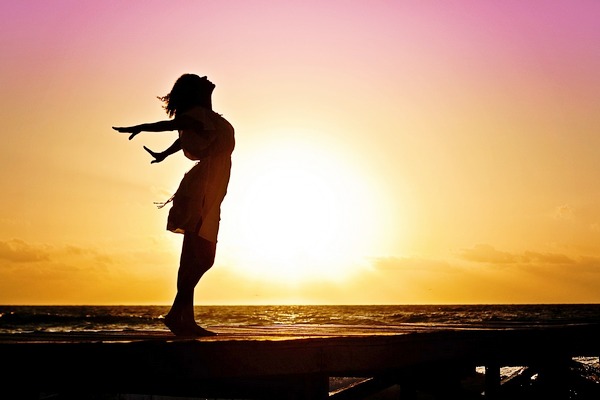 A woman stands silhouetted against a rising or setting sun, with her head throw back and arms stretched back behind her