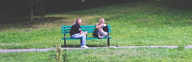 A man and a woman sit apart but facing each other and talking on a bench in a park