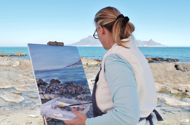 The camera looks over the shoulder of a middle-aged woman on a rocky beach painting a picture of a distant mountain