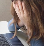 A woman sits at a table in front of her laptop with her head in hands, which cover her face 