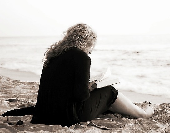 A middle-aged woman in a business suit and bare feet sits and reads a book at the beach