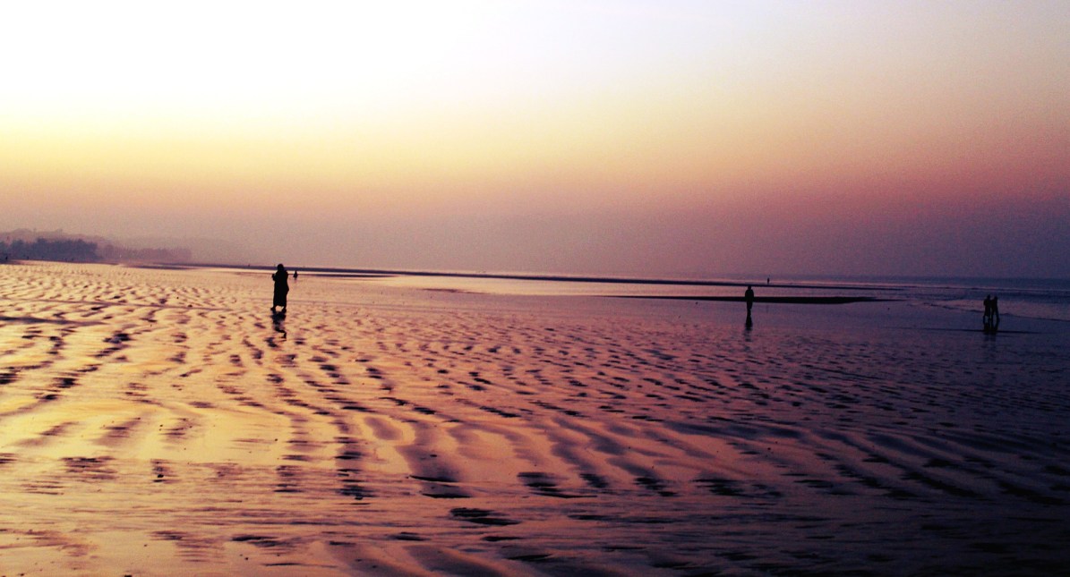 A large beach with scalloped sand patterns at dusk with a few solitary figures 