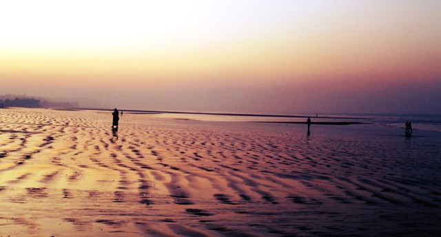 A large beach with scalloped sand patterns at dusk with a few solitary figures 