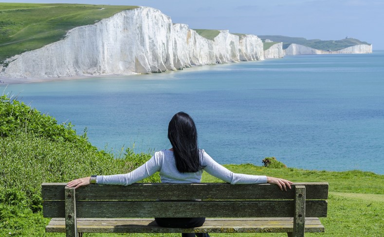 A woman with long black hair seated on a wooden bench looking over water toards the white cliffs of Dover.