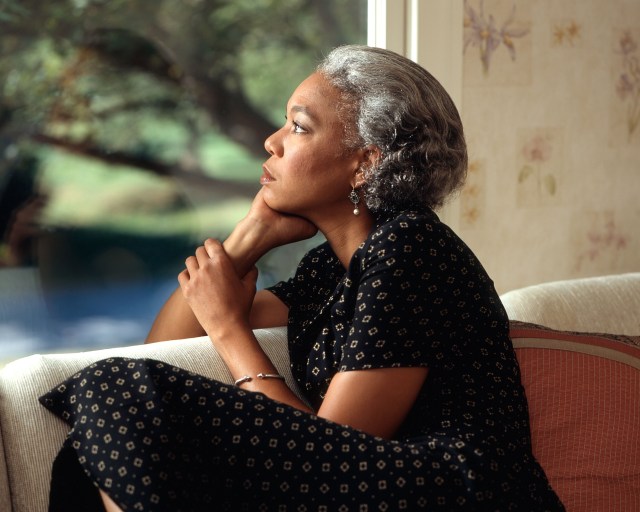 Gray-haired African American woman sits by a window, looking out and up thoughtfully.