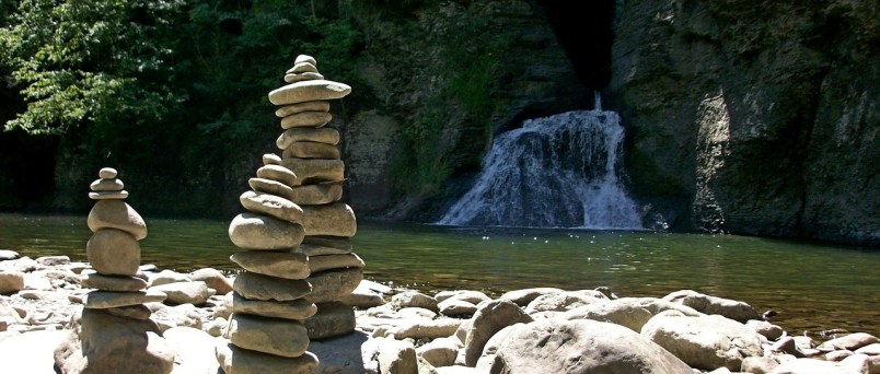 Photo by Willabee Three towers of stacked stones on a rocky river shore with a waterfall flowing down a high bank on the other side in the background