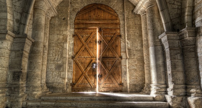 Wide stone stairs in an old stone building lead up a wooden double door. One door is ajar with light showing around its edges.