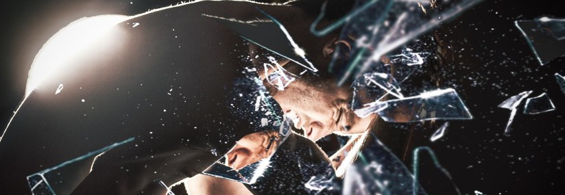 A man bending over with an expression of pain on his face is seen through a an overlay of cracked mirror shards
