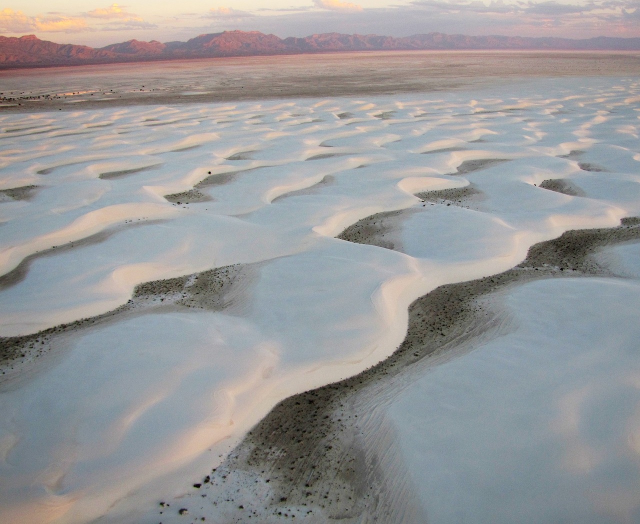 Image by skeeze (Pixabay) A large desert plain with a rippled white sand floor, edged with stony mountains in the far distance lit by a rising or setting sun.