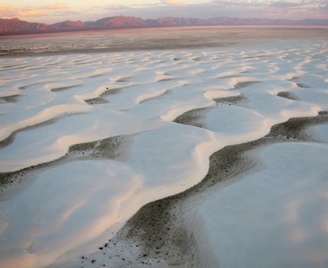 A large desert plain with a rippled white sand floor, edged with stony mountains in the far distance lit by a rising or setting sun.